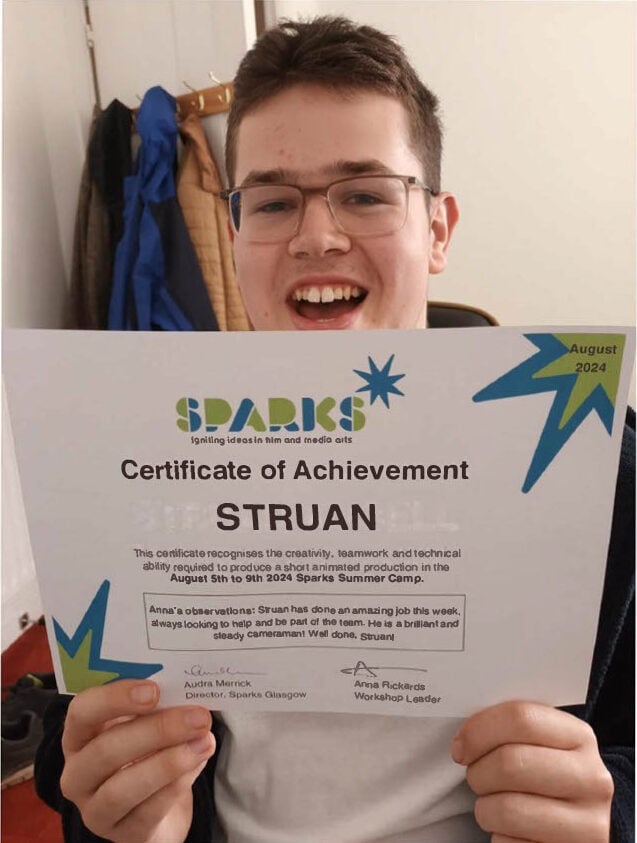 A person is holding up a “Certificate of Achievement” from Sparks Summer Camp. The certificate is awarded to someone named Struan for creativity, teamwork, and technical ability during an August 2024 film and media arts camp. The certificate includes positive comments from the workshop leaders and is decorated with colourful star graphics. Coats hang on hooks in the background.