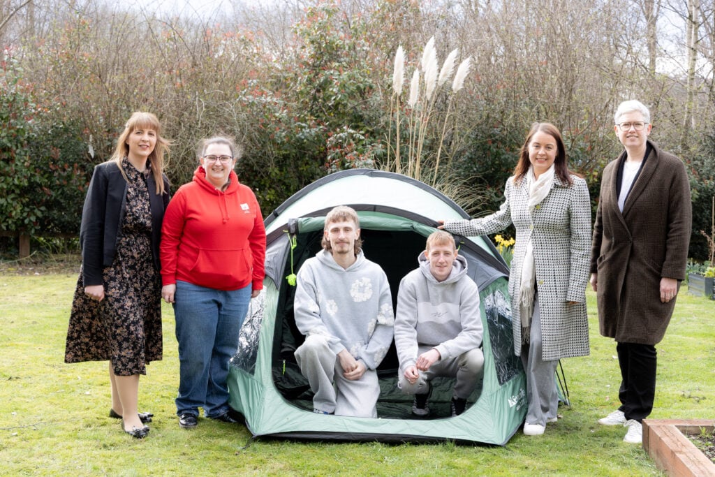 A group of six people stand and kneel around a small green tent set up on a grassy lawn. Two individuals are seated inside the tent, while the others stand on either side of it. Behind them is a backdrop of shrubs and tall plants, and the scene appears to be outdoors on a cloudy day.