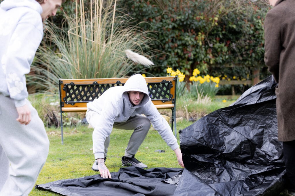 A person wearing a light grey hoodie and trainers is crouched on a grassy lawn, arranging a large black tarp on the ground. Another person in grey clothing stands to the left, partially in view, and a person in a brown coat is visible on the right. Behind them is a wooden bench with a decorative metal backrest, surrounded by tall grasses and blooming yellow daffodils. Dense greenery fills the background.