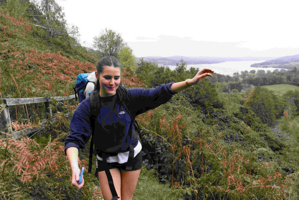 A young woman is hiking along a trail surrounded by lush greenery and ferns. She is wearing a navy blue sweatshirt and black shorts, with a backpack on her back. The landscape features rolling hills and a lake in the background, under a cloudy sky. The woman appears to be enjoying her hike, with one arm outstretched as she balances on the path.