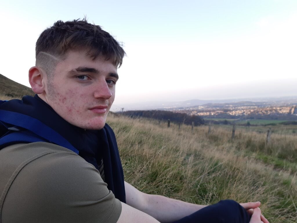A young person sits on a grassy hillside wearing outdoor clothing and a backpack, viewed from the side. The foreground shows long grass and a fence line, while the background opens onto a wide valley with buildings, fields, and distant hills under a pale sky, suggesting a rural landscape seen from higher ground.