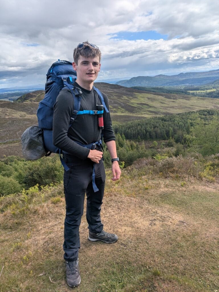 A young person stands on open hillside terrain wearing outdoor clothing and a large backpack with chest straps fastened. A water bottle is clipped to the front of the rucksack, and walking boots are visible. The landscape behind shows rolling hills, patches of woodland, and distant ridgelines under a cloudy sky, suggesting a rural upland walking environment.