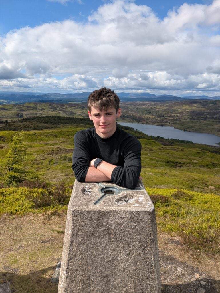 A young person stands at a hilltop trig point, resting their arms on the concrete pillar. The background shows a wide upland landscape with rolling green hills, patches of woodland, and a loch in the valley below. Distant ridgelines stretch across the horizon beneath a sky filled with large white clouds.