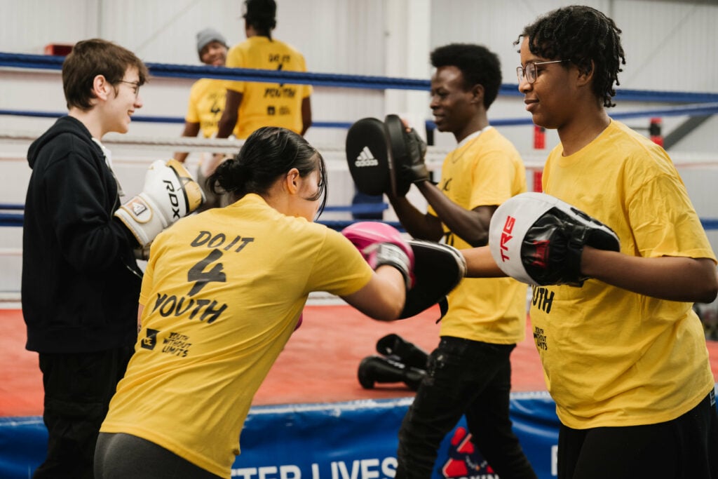 A group of young people in yellow t-shirts engaged in a boxing training session in a gym. One person is wearing pink boxing gloves and practicing punches with another, while two others are smiling and interacting nearby. The gym features a boxing ring and training equipment in the background.