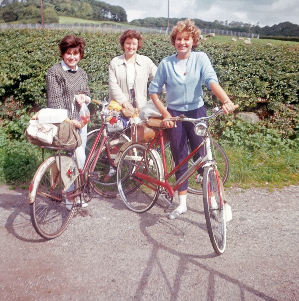 Three women stand beside their bicycles on a gravel path, surrounded by greenery. They are smiling and dressed in casual clothing, with one woman wearing a light blue sweater. Each woman has a bag or basket on her bike, suggesting they are on a leisurely outing. The background features a lush landscape with trees and fields under a partly cloudy sky.
