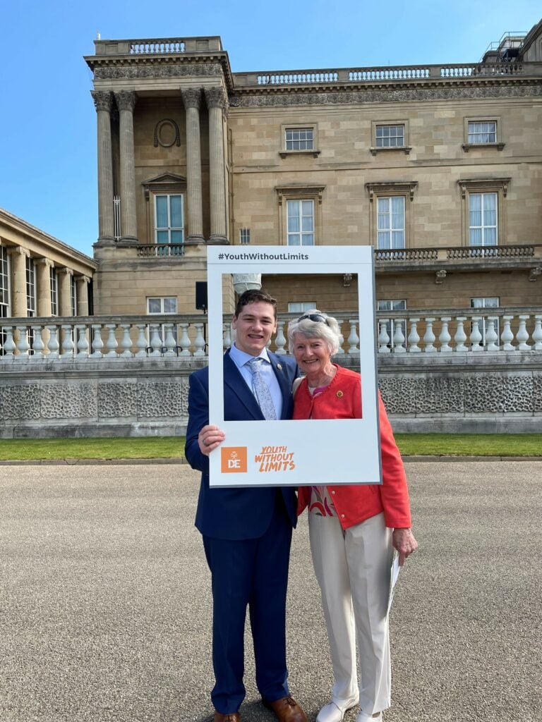 A young man and an older woman pose for a photo in a selfie frame with a "DofE Youth Without Limits" logo on. They stand in front of a grand building, with gravel underneath and a small patch of greenery. There is blue sky in the background.