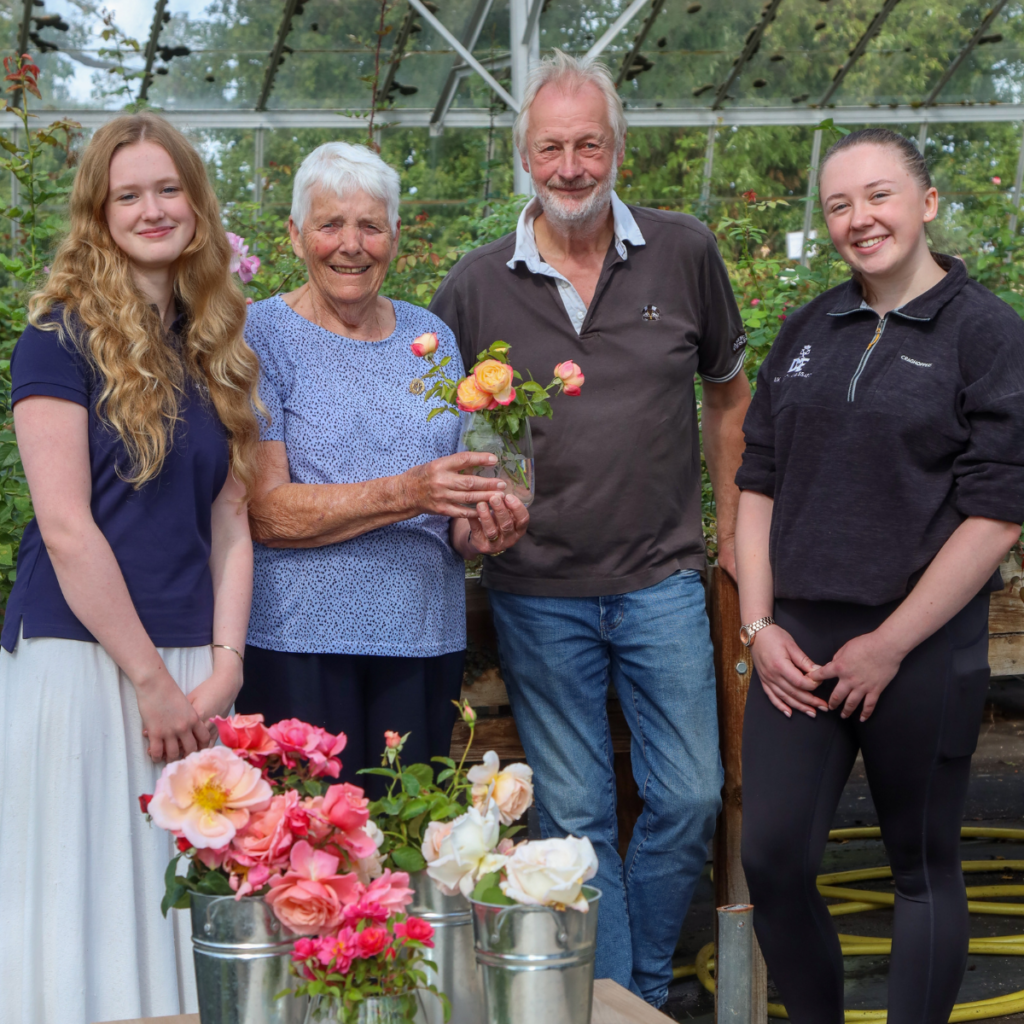 Image of a group of people standing around roses in vases in a greenhouse