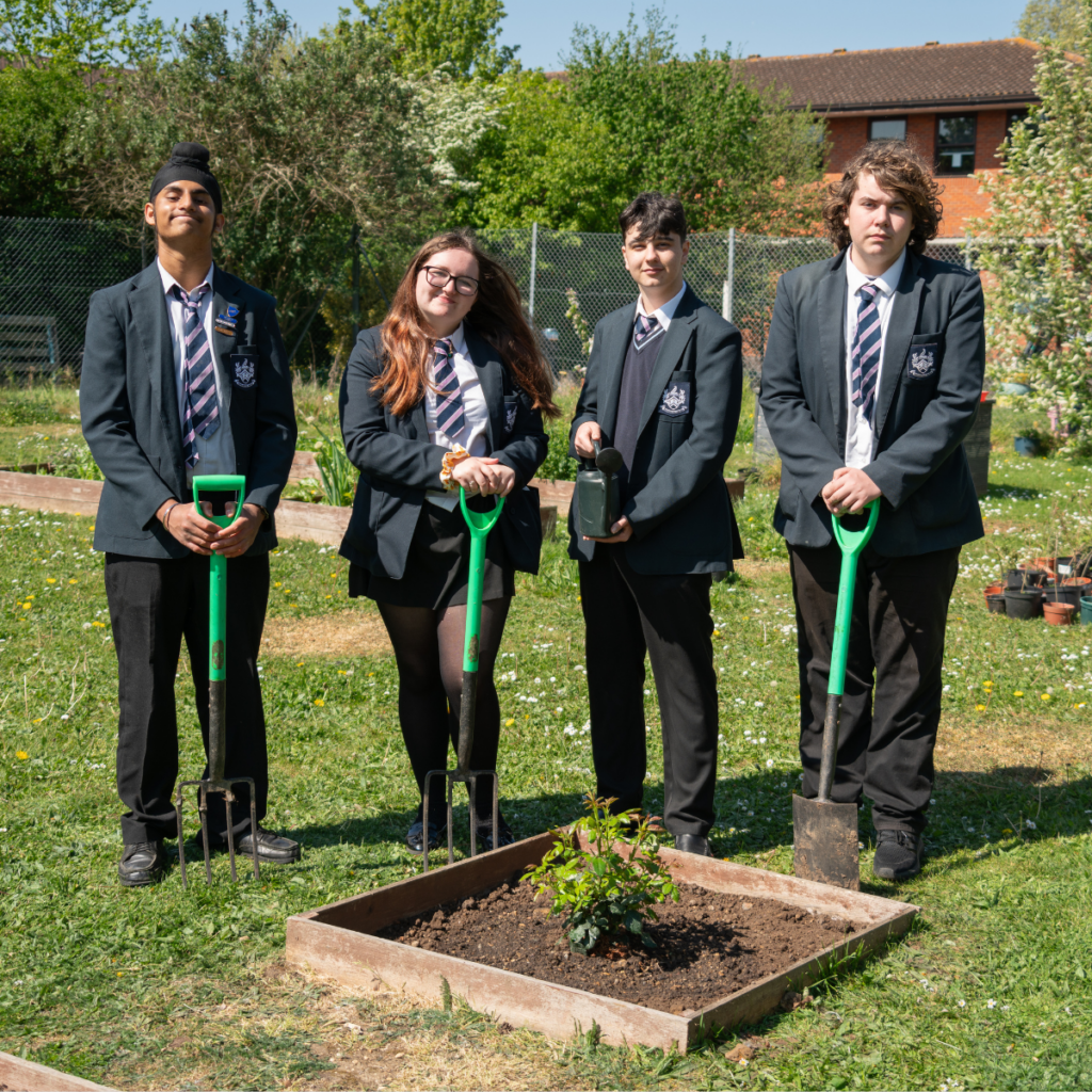 Group of young people standing outdoors on grass around a small raised garden bed with a newly planted shrub. Each person holds a spade or garden fork. A greenhouse and trees are visible behind them, with a brick building in the background under a clear blue sky.
