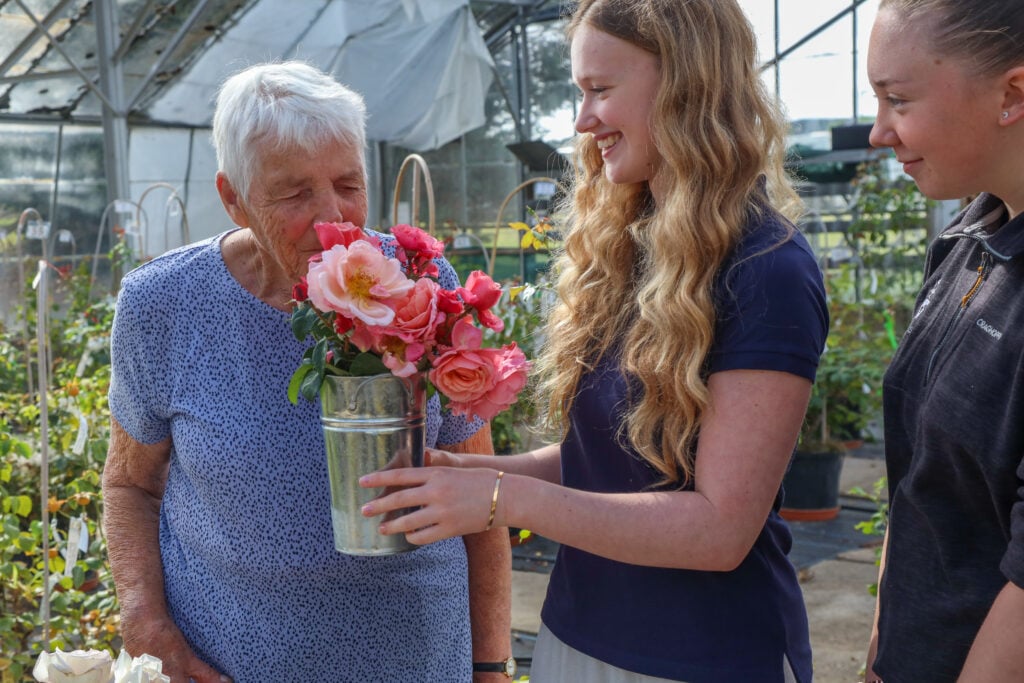 Two people standing inside a greenhouse hold a metal container filled with pink and coral roses. The flowers are being presented between them at chest height. Rows of potted plants and metal supports are visible around them, with soft natural light coming through the glass structure.