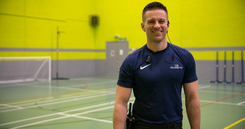 Tom in a navy blue t-shirt that says "Tom, Wellbeing PCO" standing in a sports hall with neon green walls.