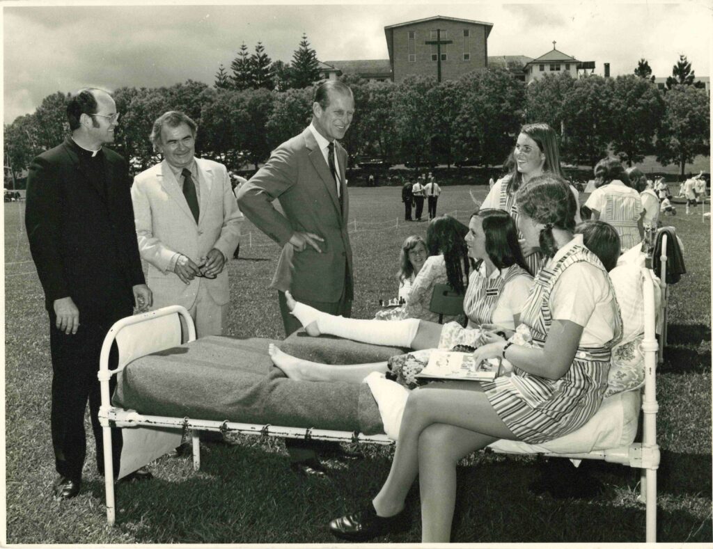 Black-and-white photograph of a group gathered on a grass field. In the centre, HRH The Duke of Edinburgh, Prince Philip, stands with one hand on their hip, speaking to several young people seated on and around a metal-framed bed placed outdoors. One young person lies on the bed with a leg in a plaster cast, while others sit nearby wearing striped uniforms. To the left, two adults in formal clothing, including one in clerical dress, stand observing. In the background, more people are scattered across the field, with a large building featuring a prominent cross and a row of trees behind it.