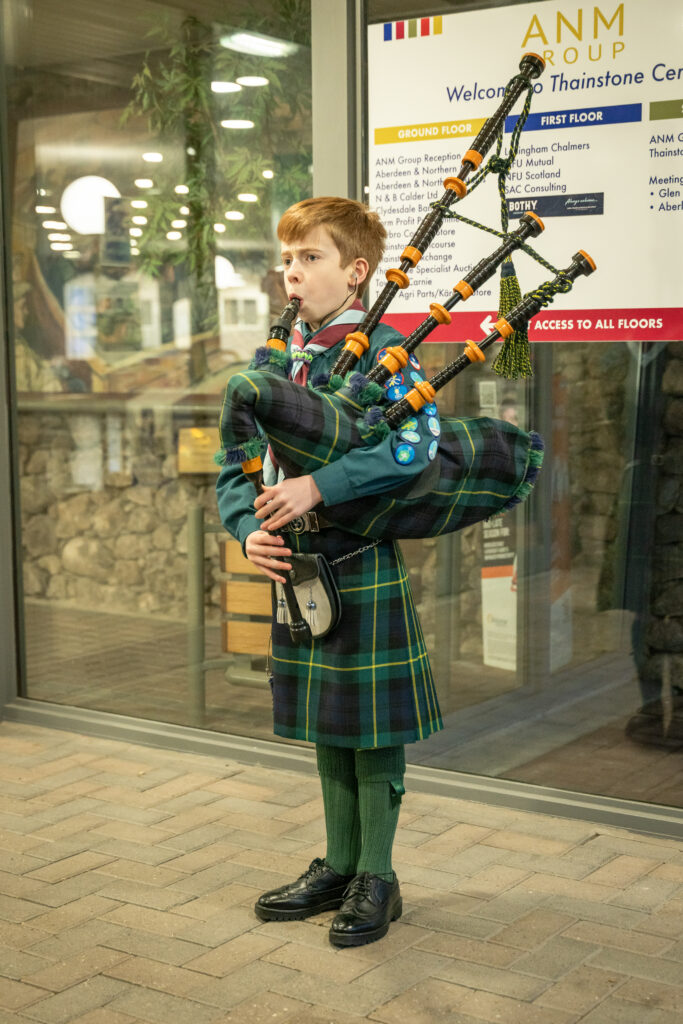 A young bagpiper in a tartan kilt stands indoors playing the bagpipes. The piper wears a uniform with several badges and stands in front of glass windows and an information sign at the Thainstone Centre. The bagpipes feature orange decorative accents and green tassels.
