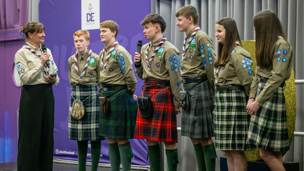 A group of six young people in Scout uniforms and tartan kilts stand on a stage at a DofE Scotland event. One participant speaks into a microphone while another person holding a microphone stands opposite them. A purple DofE Scotland display and grey stage curtains form the backdrop.