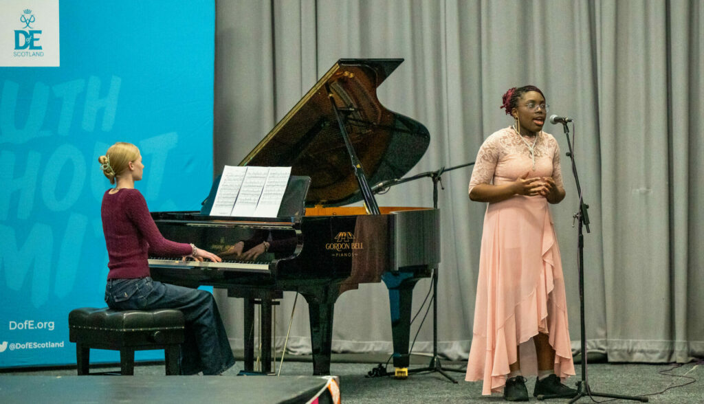 A performer stands beside a microphone on stage while another performer plays a grand piano. A blue DofE Scotland banner stands to one side, with grey curtains forming the backdrop. The stage is decorated with colourful bunting, and sheet music is visible on the piano.