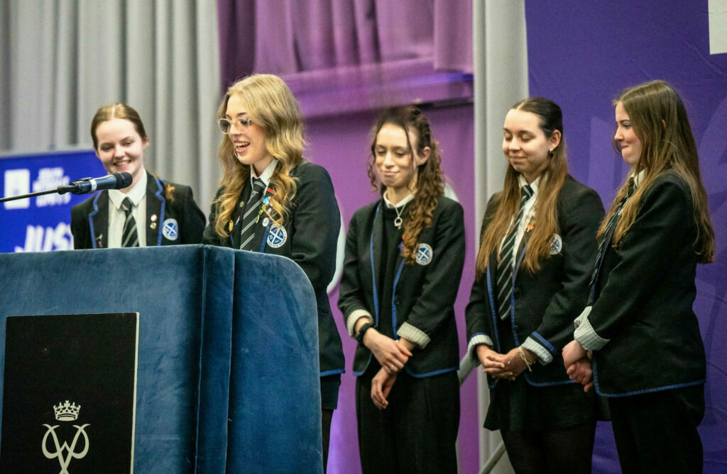 Five students in school uniform stand on a stage at a DofE Scotland event. One student speaks at a lectern with a microphone while the others stand nearby. The lectern displays the Duke of Edinburgh's Award crest, with purple DofE Scotland branding and grey stage curtains in the background.