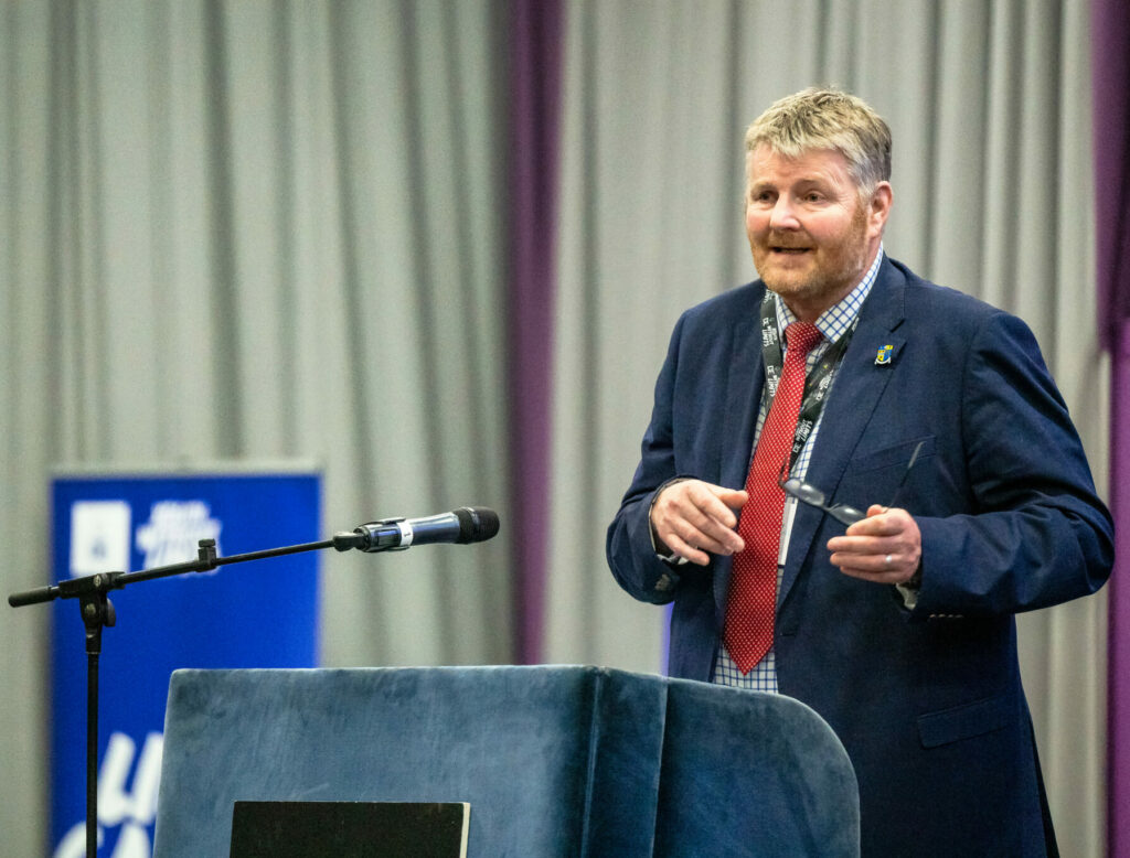 A speaker stands behind a lectern on a stage at a DofE Scotland event, addressing an audience. The lectern displays the Duke of Edinburgh's Award crest, with a microphone positioned nearby. Grey curtains and DofE Scotland branding form the backdrop.