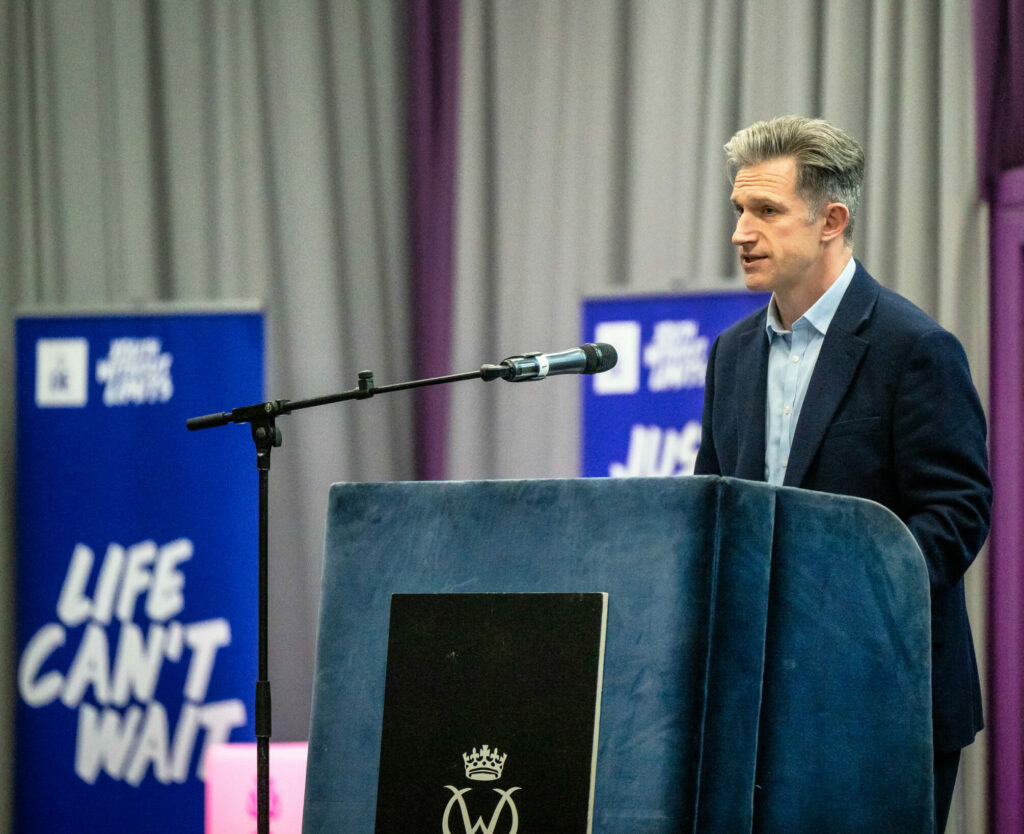 A person stands behind a lectern speaking at a DofE Scotland event. Two blue banners displaying the DofE slogan "Life Can't Wait" are positioned behind the stage, with grey curtains in the background. A microphone is mounted beside the lectern, which features the Duke of Edinburgh's Award crest.