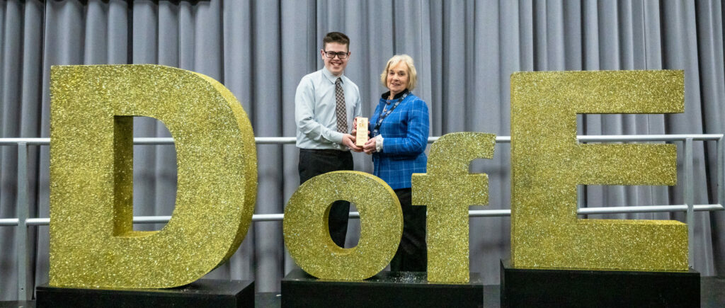 Two people stand on a stage behind large glittery gold letters spelling "DofE", holding an award plaque together. Grey curtains form the backdrop, with the oversized letters mounted on black bases across the front of the stage.