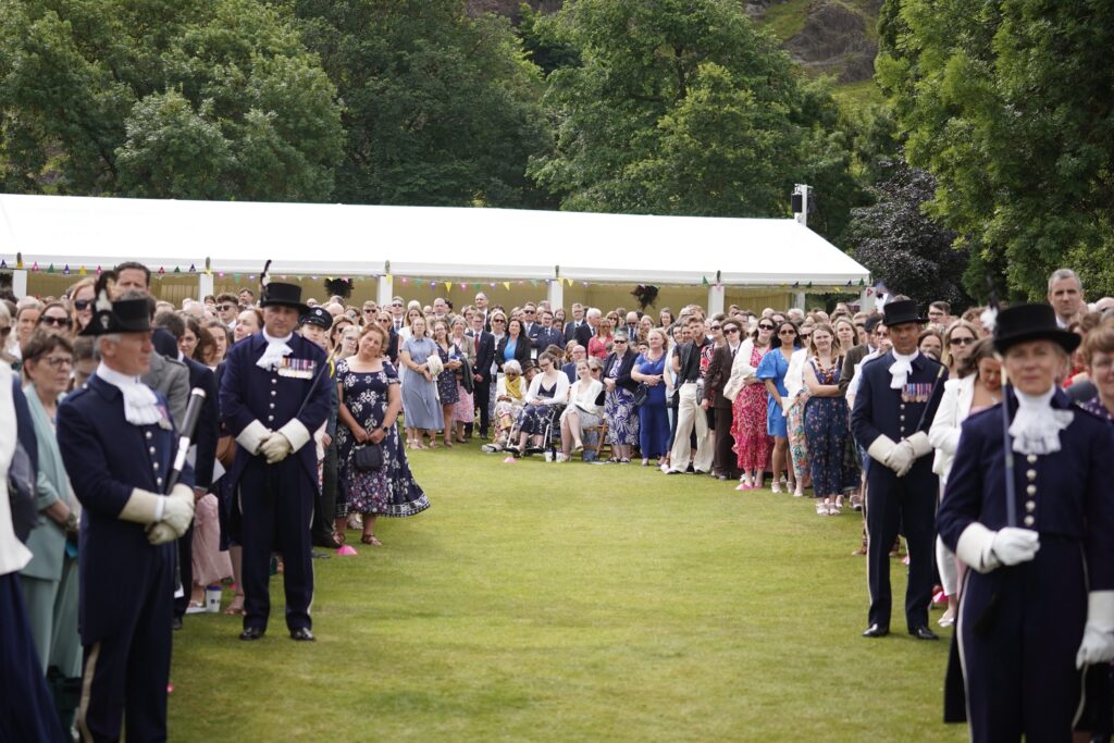 Large crowd gathered on a lawn at the DofE Gold Award Celebration at the Palace of Holyroodhouse. Two rows of ceremonial attendants in dark uniforms and top hats stand on either side of a grass walkway, with attendees lined behind them. A large white marquee and leafy trees form the backdrop on a bright summer day.