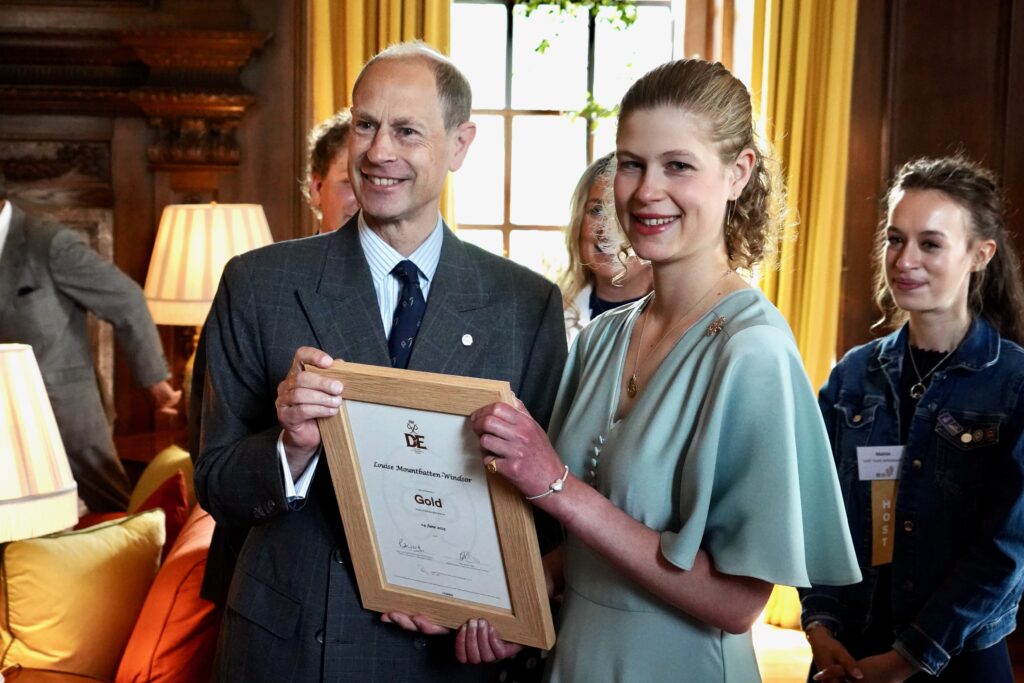 Lady Louise Mountbatten-Windsor holding a framed DofE Gold Award certificate alongside another person in a formal suit during an indoor presentation at a wood-panelled venue. Several attendees stand nearby, and large windows with gold curtains and table lamps can be seen in the background.