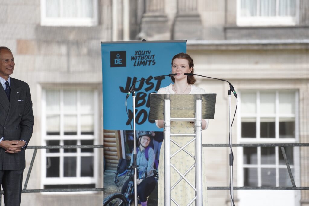 Isla stands at a lectern on an outdoor stage at the Palace of Holyroodhouse, speaking into two microphones. A DofE banner displaying the slogan “Youth Without Limits” stands behind the lectern, while attendees and the palace building can be seen in the background.