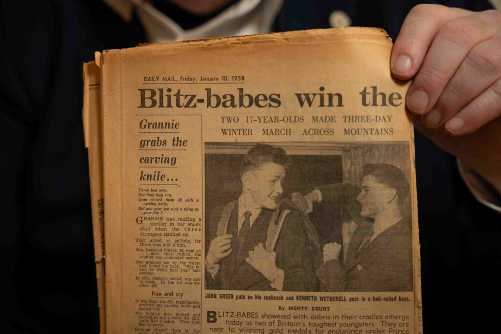 John Green holding an old newspaper titled "Daily Mail" from January 10, 1958. The headline reads "Blitz-babes win the" with a subheading about two 12-year-olds completing a three-day winter march across mountains. The article features a photo of two young boys, one holding an axe, engaged in conversation. The newspaper appears worn and yellowed with age.