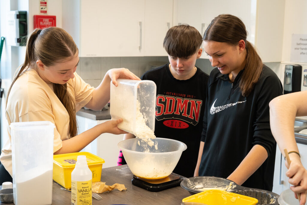 Three young people in the kitchen looking at a bowl on a scale as one of them pours flour in.