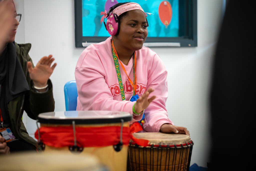 A young person wearing a pink jumper and pink ear defenders playing a djembe drum.