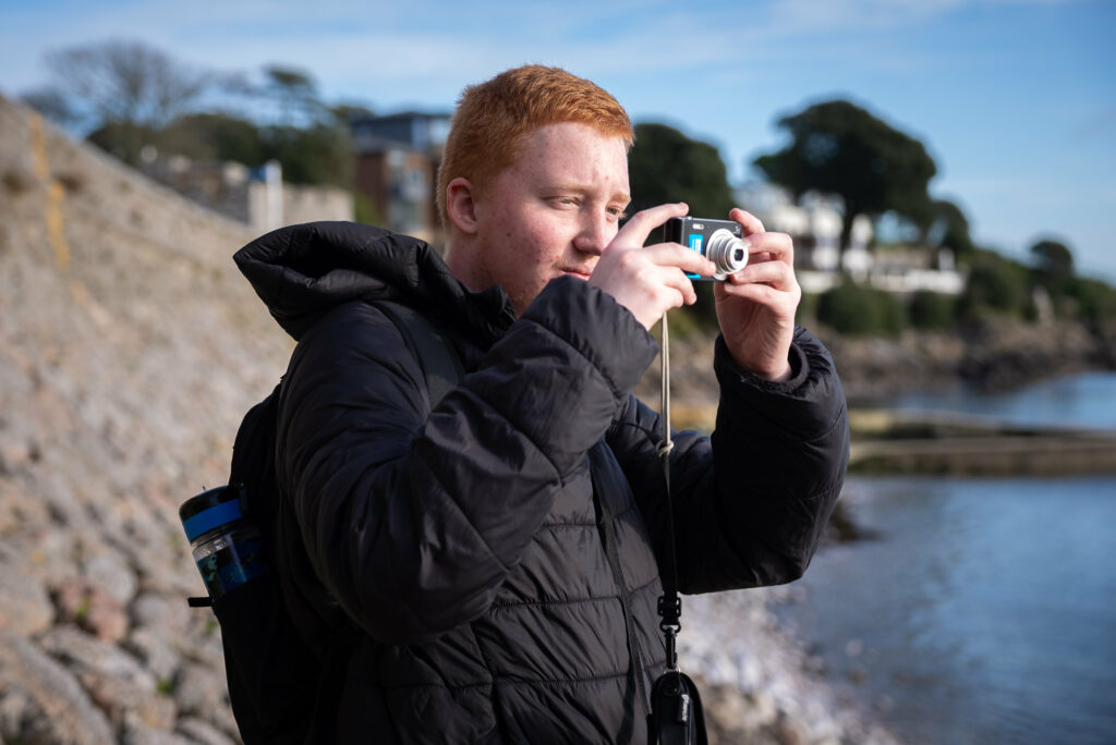 A young person in a black puffer jacket standing by the shore holding a digital camera.