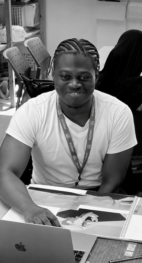 Umar Dumbuya wearing a white T-shirt and a student lanyard sits at a worktable with an open laptop and a large design portfolio. One hand rests on the portfolio while shelves, chairs and studio equipment are visible in the background. The black-and-white photo is taken indoors in a creative workshop or classroom setting.