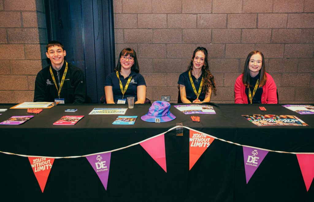 Four DofE Youth Ambassadors sit behind an information table at an indoor event venue. The table is covered with DofE Scotland and Youth Without Limits bunting and displays leaflets, posters, pens, a clipboard, and a purple DofE bucket hat. The volunteers wear DofE-branded clothing and lanyards, with a brick wall backdrop behind the stand. Visible materials promote DofE opportunities and activities for young people.