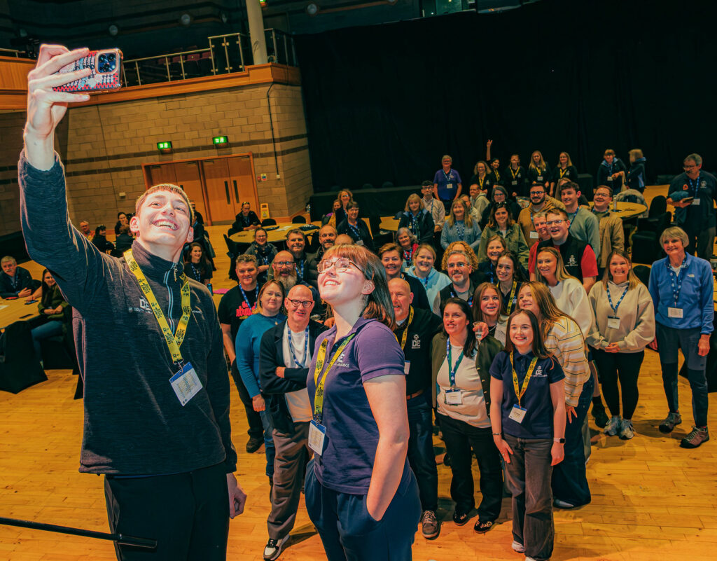 A large group of attendees gathers in a conference hall for a group photo during a DofE event. In the foreground, two DofE Youth Ambassadors wearing lanyards stand on a stage taking a selfie with a smartphone, while dozens of participants and staff members pose behind them. Round tables are arranged throughout the venue, and the spacious hall features a wooden floor, tiered seating, and event activities taking place in the background.