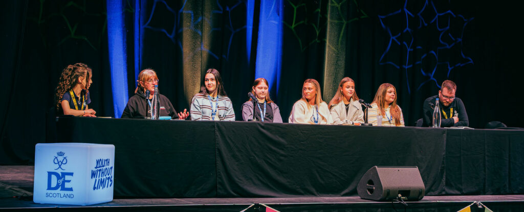 A panel discussion takes place on stage at a DofE Scotland event, with seven panellists seated behind a long table facing an audience. Microphones, water bottles and event materials are arranged on the table, while DofE Scotland and Youth Without Limits branding is displayed on illuminated cubes and bunting across the front of the stage. Blue and green lighting highlights the backdrop curtains as attendees watch from round tables in the conference hall.