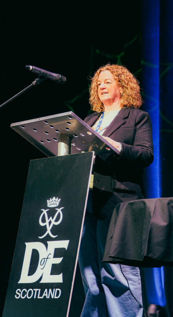 A speaker stands at a podium displaying the DofE Scotland logo, delivering a presentation on a stage in a large conference venue. A microphone is positioned beside the lectern, and blue stage lighting illuminates the dark backdrop curtains. A table with presentation equipment sits nearby, while DofE branding is prominently featured at the front of the stage.