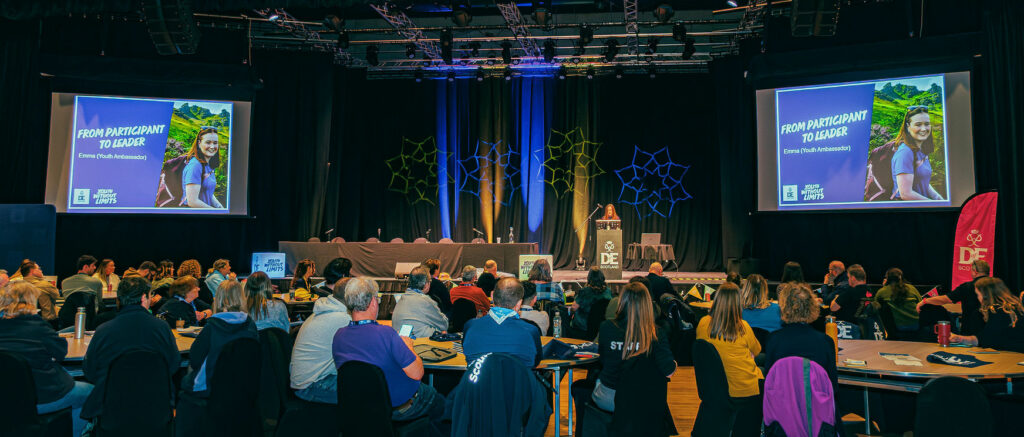 Attendees sit at round tables in a large conference hall while watching a presentation on a stage at a DofE Scotland event. Two large screens display a slide titled “From Participant to Leader,” featuring a DofE Youth Ambassador. The stage is lit with blue and green lighting and includes DofE Scotland and Youth Without Limits branding, while participants take notes and listen from tables arranged across the venue.