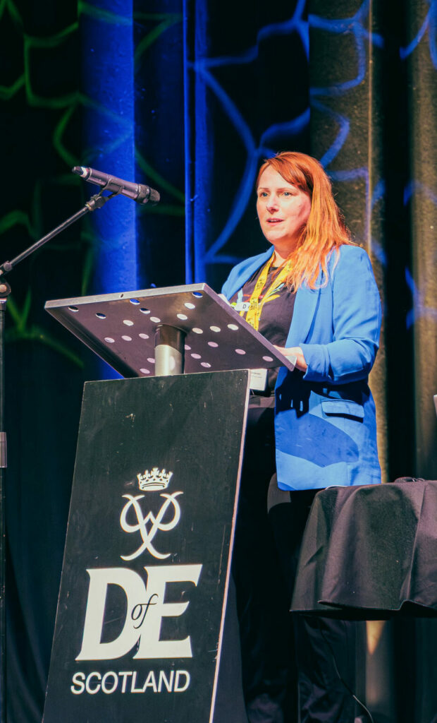A speaker stands behind a podium displaying the DofE Scotland logo, addressing an audience from a stage at an indoor event. Blue and green stage lighting illuminates the backdrop curtains, while a microphone stand and a table with a laptop are positioned nearby. DofE and Youth Without Limits branding is visible around the stage area as the presentation takes place in a conference-style venue.