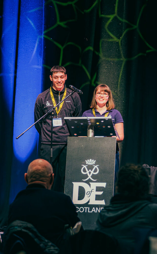 Two DofE Youth Ambassadors stand at a podium displaying the DofE Scotland logo, presenting to an audience from a conference stage. A microphone and lectern are positioned in front of them, with a laptop on a nearby table. Blue and green stage lighting illuminates the backdrop curtains, while attendees seated in the foreground watch the presentation.