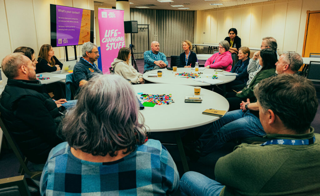 Participants sit around three circular tables during a DofE workshop in a conference room. Colourful building bricks are spread across the tables as part of a group activity, while a facilitator stands beside the group. A presentation screen displays discussion prompts and a DofE banner reading “Life-Changing Stuff” stands nearby. Notebooks, drinks, and laptops are visible as participants take part in a collaborative discussion.