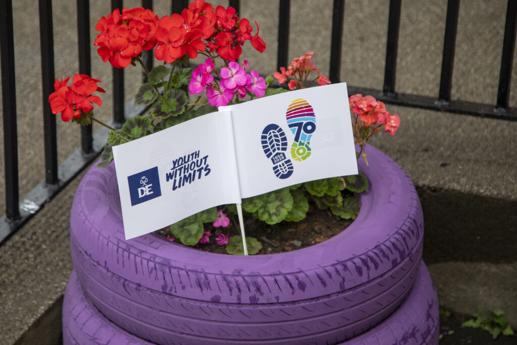 A close-up of a flower planter made from stacked purple-painted tyres, filled with bright red and pink flowering plants. Two small flags are placed among the flowers, displaying the DofE logo, the text “Youth Without Limits,” and a colourful 70th anniversary footprint design. A stone wall and black metal railings form the background. The image highlights DofE branding integrated into a colourful outdoor floral display.