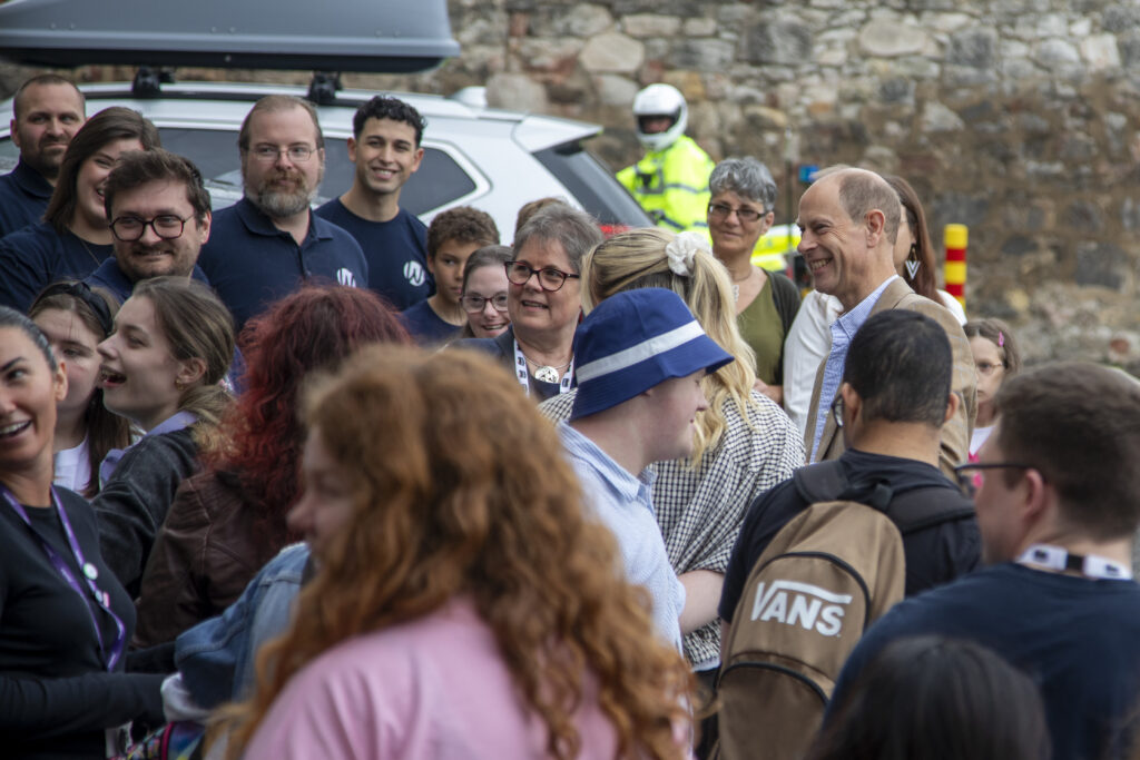 A large group of people gather outdoors in a car park beside a stone wall and parked vehicles. Near the centre of the crowd, a person in a tan checked blazer speaks with attendees, while others stand nearby wearing lanyards and casual clothing. The scene includes people of different ages, with several participants facing toward the central conversation. A high-visibility motorcycle rider can be seen in the background, and the image captures a busy community event atmosphere with many attendees interacting and listening.
