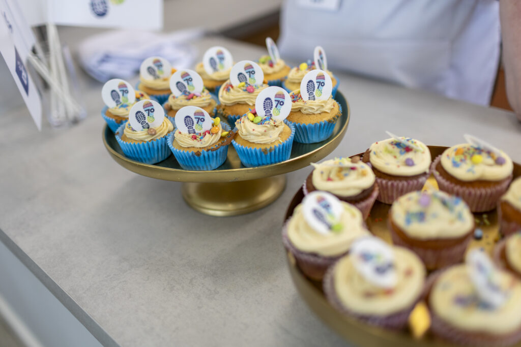 A close-up view of decorated cupcakes displayed on two cake stands on a countertop. The cupcakes are topped with swirls of pale icing, colourful sprinkles, and edible decorations featuring a footprint logo and the number “70”. Cupcakes in bright blue and pink paper cases are arranged neatly on gold-coloured stands. The background is softly blurred, with part of a white apron and event materials visible behind the display.