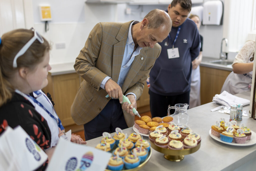 A group of people gather around a countertop in a kitchen or catering space while decorating cupcakes. His Royal Highness, The Duke of Edinburgh uses a piping bag to add icing to plain cupcakes, while others wearing event lanyards watch nearby. Decorated cupcakes topped with colourful sprinkles and circular “70” logo decorations are displayed on cake stands and plates in the foreground. Kitchen units, a sink, and baking supplies are visible in the background, creating a hands-on celebration and baking activity setting.