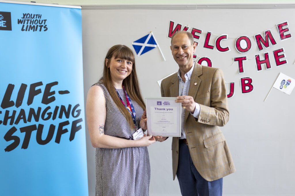 Two people stand together indoors holding a framed certificate during a presentation. One person wears an event lanyard and patterned dress, while the other wears a tan checked blazer. Behind them is a blue Duke of Edinburgh’s Award banner displaying the text “Youth Without Limits” and “Life-Changing Stuff”, alongside a wall display with a welcome message, a Scottish flag, and DofE 70th anniversary decorations. Both individuals face the camera while presenting the certificate in a bright community venue.