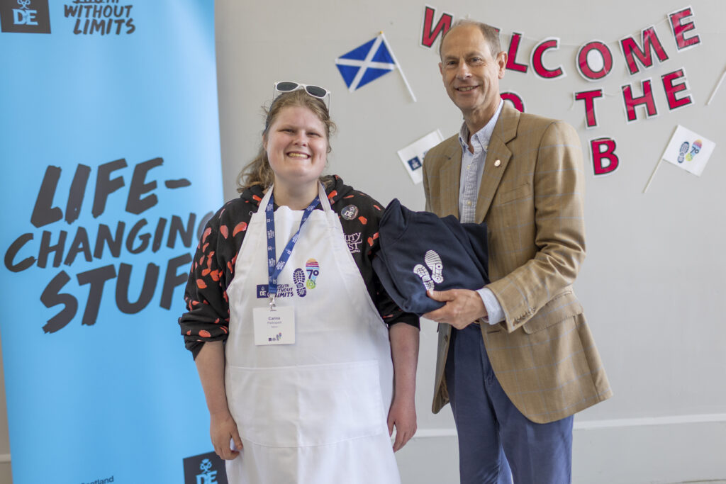 Two people stand indoors in front of a Duke of Edinburgh’s Award display during a presentation event. One person wears a white apron featuring the DofE 70th anniversary logo and an event lanyard, while the other wears a tan checked blazer and holds a folded navy garment with a DofE footprint design. Behind them are a blue “Youth Without Limits” and “Life-Changing Stuff” banner, a Scottish flag, and a welcome display with DofE anniversary decorations. Both individuals face the camera in a bright community venue setting.