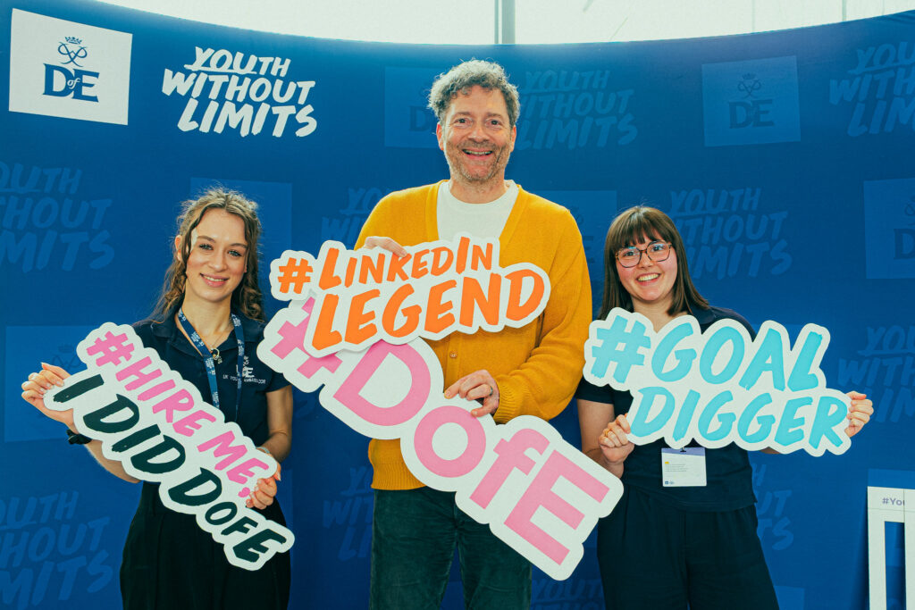 Three event attendees pose in front of a blue Youth Without Limits and DofE-branded backdrop, holding oversized hashtag props reading “#LinkedInLegend,” “#DofE,” “#HireMeDofE,” and “#GoalDigger.” All three wear event lanyards and stand in the event photo area, with Youth Without Limits and DofE logos visible behind them.