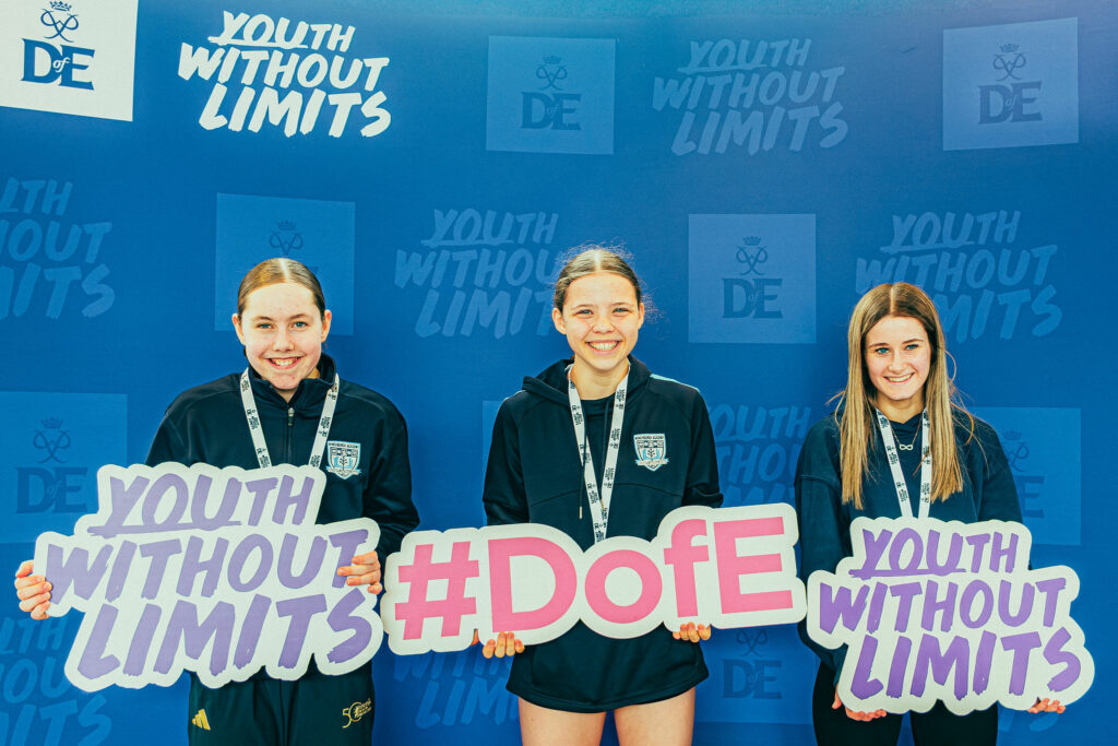 Three young attendees pose in front of a blue Youth Without Limits and DofE-branded backdrop, holding large signs that read “Youth Without Limits” and “#DofE.” All three wear event lanyards and stand in the event photo area, with Youth Without Limits and DofE logos visible throughout the background.