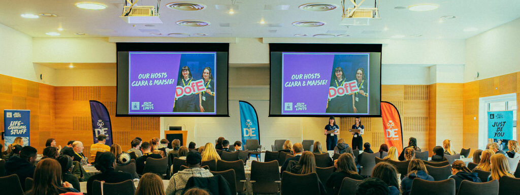 A large audience sits at round tables in a conference hall, facing a stage with two presenters. Two large screens display a slide introducing “Our Hosts Ciara & Maisie” with DofE and Youth Without Limits branding. DofE Scotland banners stand on either side of the stage, and attendees watch the event from across the room.