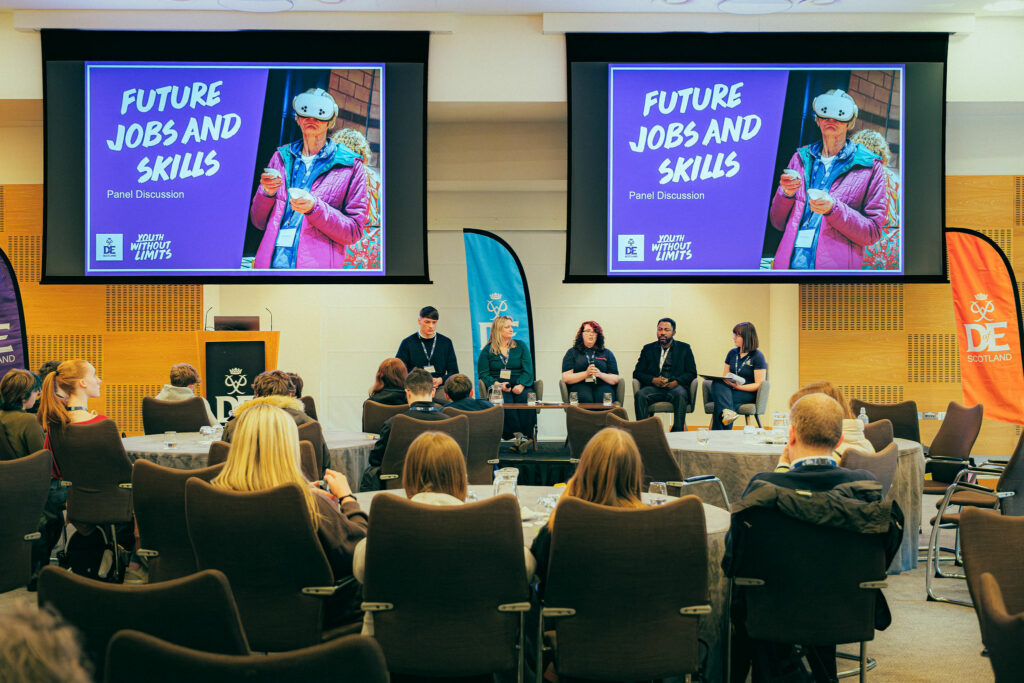 Audience seated at round tables watching a panel discussion at a conference. Five speakers sit on stage beneath two large screens displaying a slide titled “Future Jobs and Skills – Panel Discussion,” alongside an image of a person using a virtual reality headset. DofE Scotland and Youth Without Limits branding is visible on the screens and event banners.