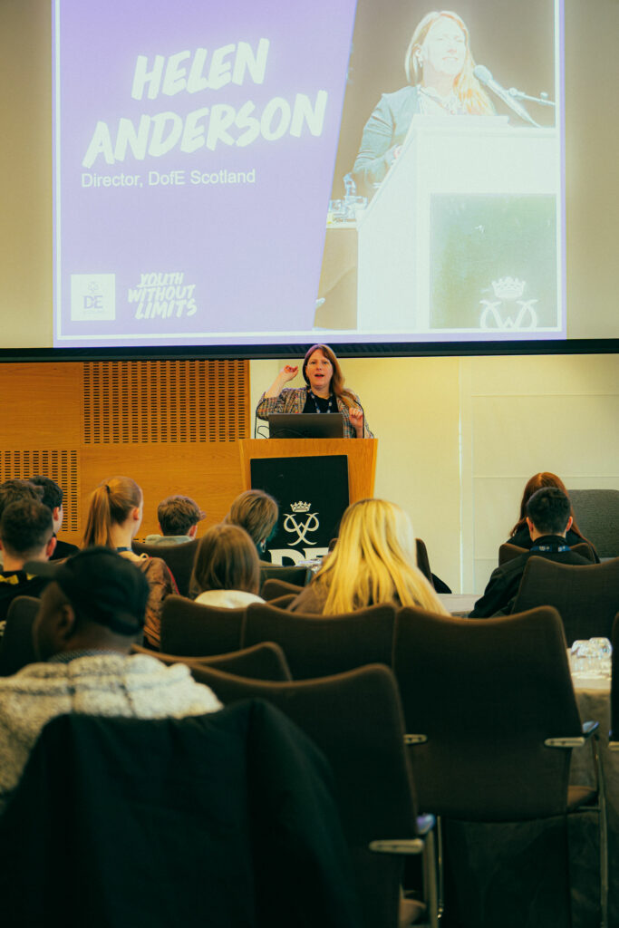 A speaker stands at a DofE-branded podium addressing an audience in a conference hall. A large screen behind the podium displays a presentation slide reading “Helen Anderson, Director, DofE Scotland,” alongside a photo of the speaker at the lectern. Attendees are seated facing the stage, listening to the presentation.