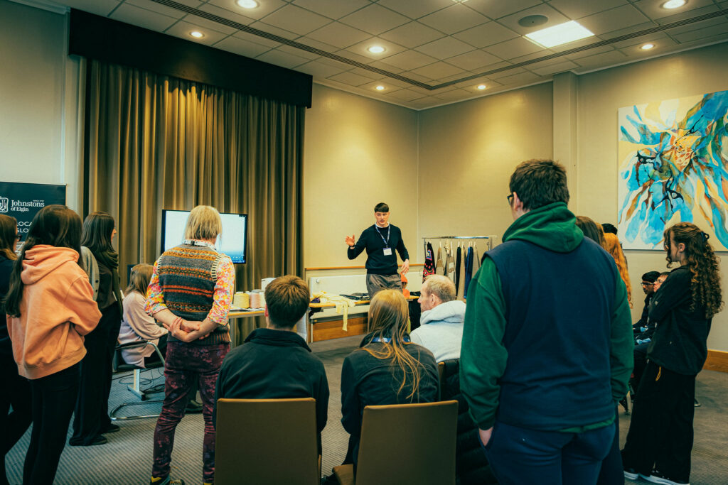 A workshop facilitator stands at the front of a room delivering an interactive session to a group of young people and adults gathered in a circle. Some participants are seated while others stand around the room, facing the facilitator. A table with materials, a display screen and a clothing rack are visible, suggesting a hands-on careers or skills-based workshop.